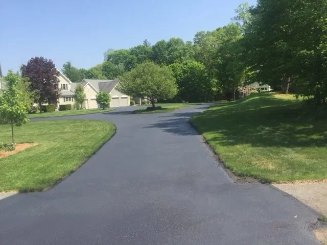 A freshly sealcoated residential driveway with a smooth, deep black finish, bordered by green lawns and mature trees in Boxborough, MA.